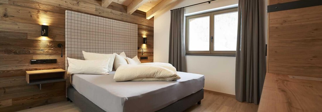 A modern bedroom with wood paneling and simple furniture. Large windows let in plenty of daylight and create a bright atmosphere. - The Peak Sölden - Sölden