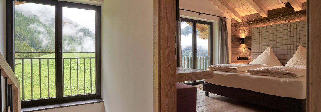 A cozy room with a double bed and wooden ceilings. Through the window, you can see a green landscape with foggy mountains. - The Peak Sölden - Sölden