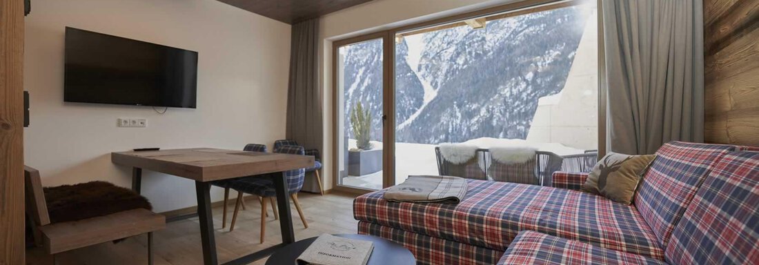 A modern living room with a cozy sofa and a dining table. Large windows offer a view of the mountains. - The Peak Sölden - Sölden