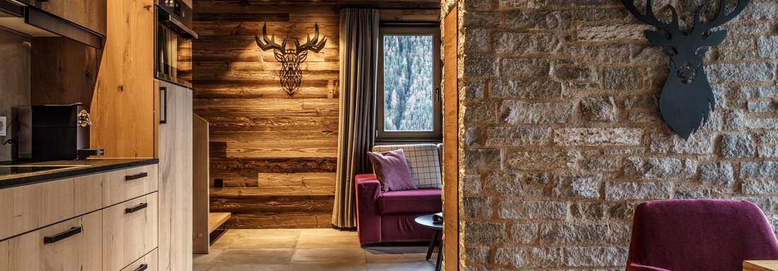 A cozy room with wooden paneling and modern furniture. A kitchenette is visible, as well as a special wall decoration with a stag's antler. - The Peak Sölden - Sölden