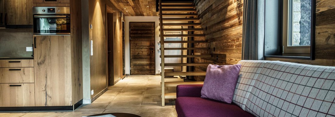 A modern living room with wood paneling and a cozy seating area. In the background, a staircase and a kitchen are visible. - The Peak Sölden - Sölden