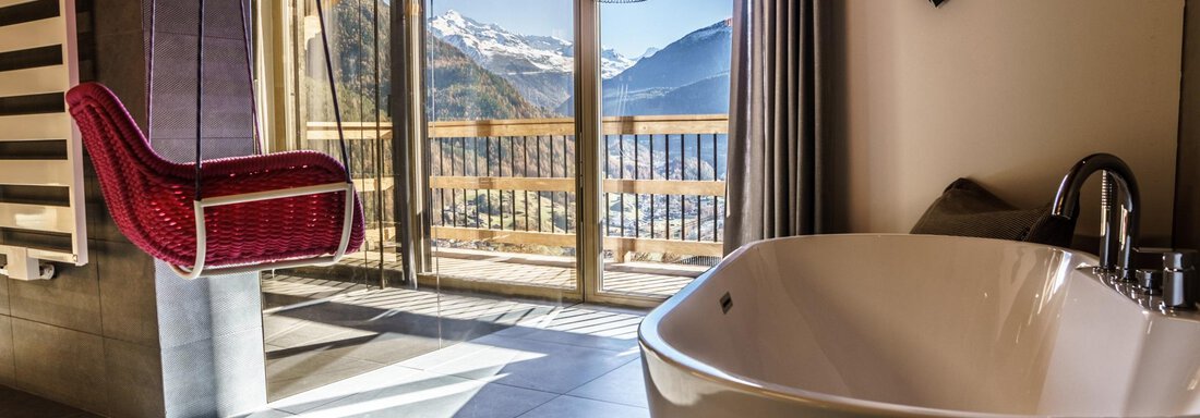 A modern bathroom with a freestanding bathtub and a cozy rocking chair. Large windows offer a stunning view of the mountains. - The Peak Sölden - Sölden