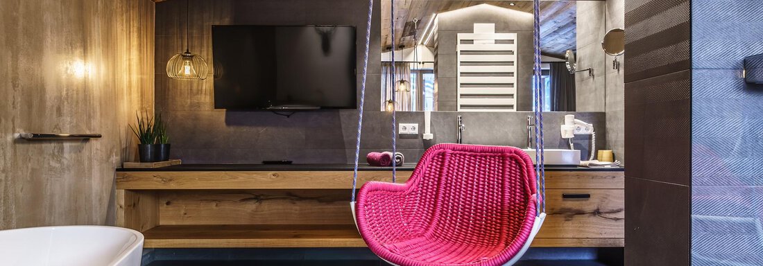 A modern bathroom with a swinging chair in pink. The decor includes wood and stone elements as well as a large television. - The Peak Sölden - Sölden