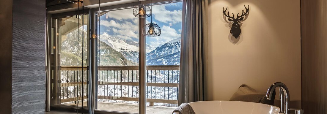 A modern bathroom with a freestanding bathtub and large windows that offer a view of the mountains. The warm wooden elements create a cozy atmosphere. - The Peak Sölden - Sölden