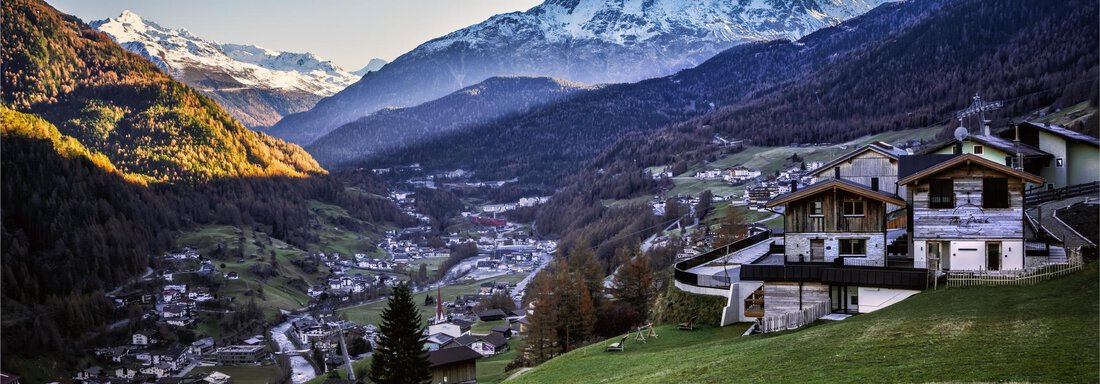 A picturesque mountain landscape with snow-capped peaks and a serene valley. In the foreground, some traditional huts can be seen. - The Peak Sölden - Sölden