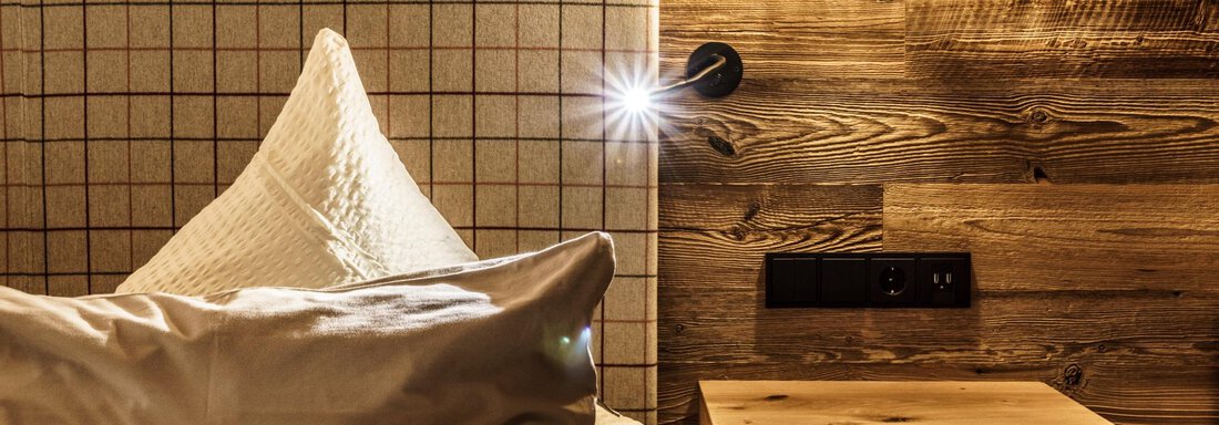 A cozy bedroom with a wooden panel and a nightstand. The bed is decorated with soft pillows and a blanket. - The Peak Sölden - Sölden