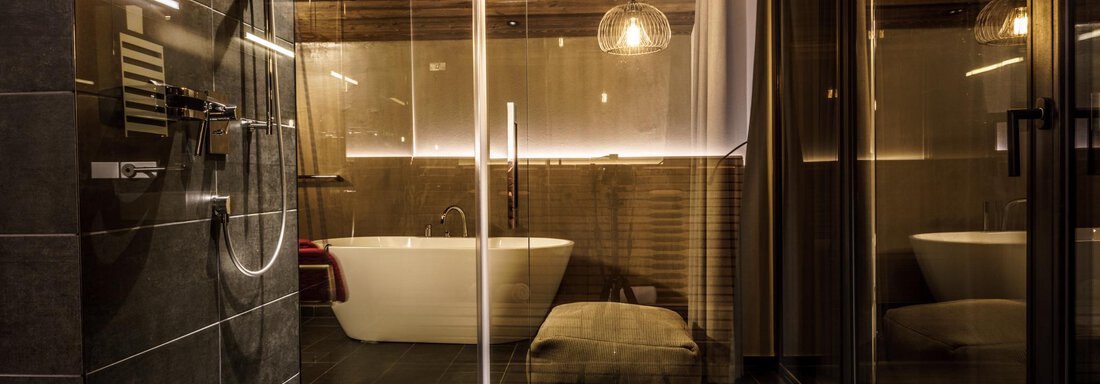 A modern bathroom with a freestanding bathtub and a glass shower. The wooden ceiling and warm lights create a cozy atmosphere. - The Peak Sölden - Sölden