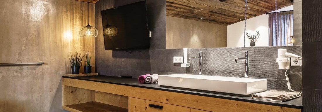 A modern bathroom with wooden details and a large mirror. On the wooden shelf, there are plants and towels, and the lighting creates a warm atmosphere. - The Peak Sölden - Sölden