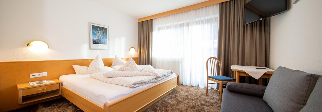 A cozy hotel room with a double bed and a gray sofa. There is a television and large windows that let in plenty of natural light. - Toni's Ferienheim - Sölden