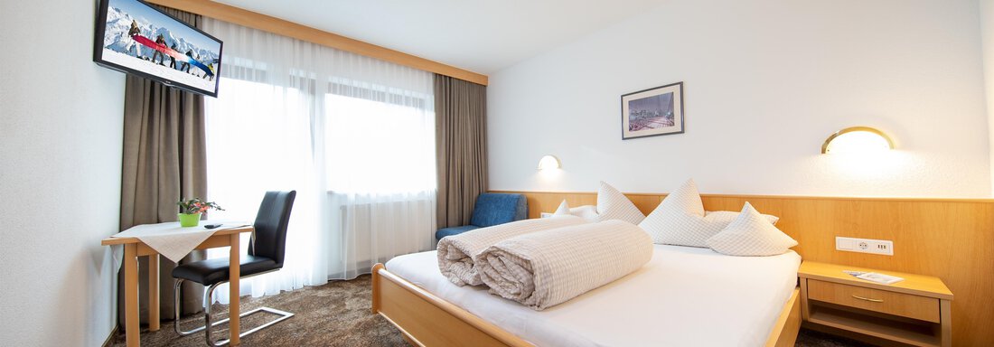 A bright hotel room with a double bed and modern furniture. There is a television, a table, and large windows with curtains. - Toni's Ferienheim - Sölden