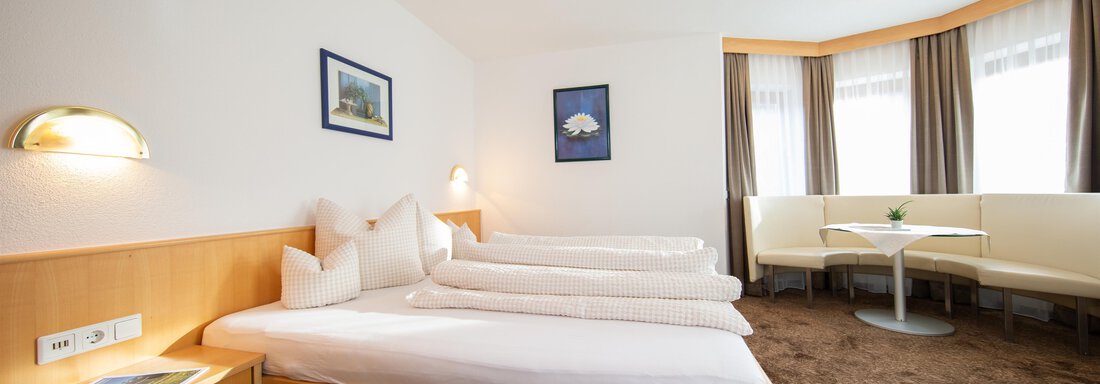 A bright hotel room with two single beds and a cozy seating area. The windows are adorned with curtains and the walls are decorated with pictures. - Toni's Ferienheim - Sölden