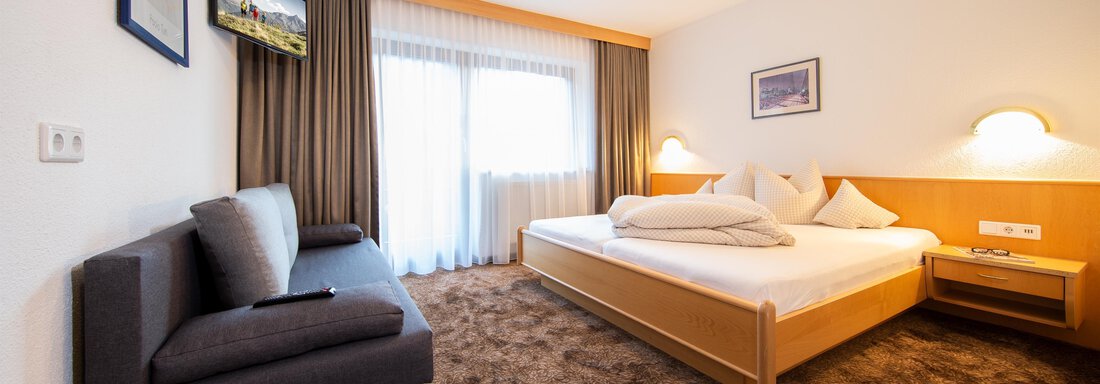 A modern hotel room with a double bed, a seating area, and a flat-screen TV. Large windows provide plenty of daylight. - Toni's Ferienheim - Sölden