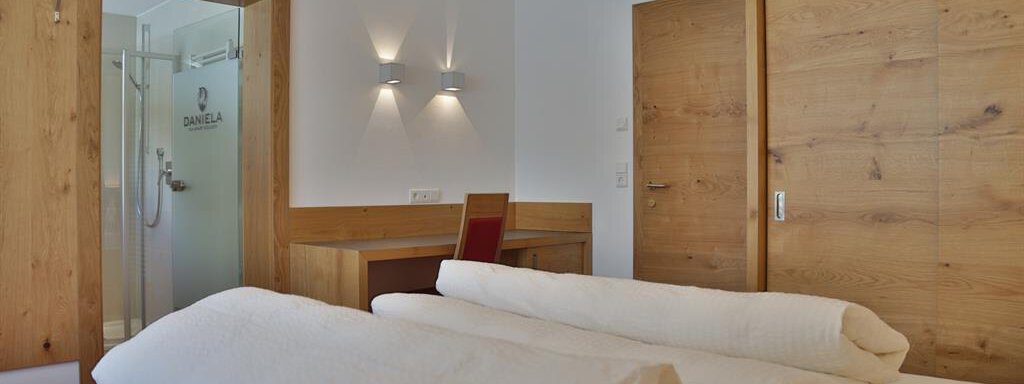 A modern hotel room with bright walls and wooden details. In the foreground are two unmade beds and in the background, there is a table and a bathroom. - Top Apart Daniela - Sölden
