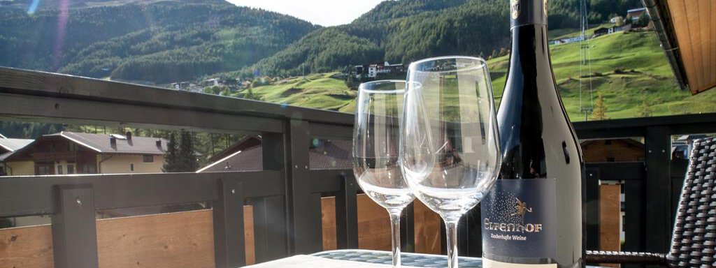 A table on a balcony overlooking green mountains. On the table are two glasses and a bottle of wine. - Top Apart Daniela - Sölden