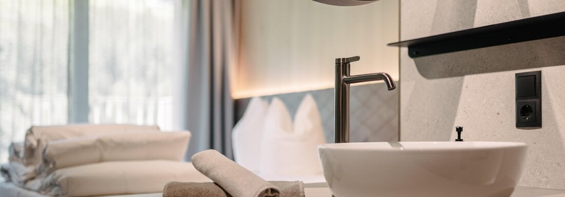 A modern bathroom with a round mirror and a sink. In the background, a comfortable bed and fresh bed linen are visible. - VIBES Sölden - your alpine homebase - Sölden