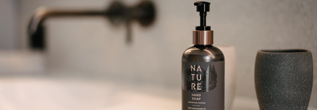 An elegant small bottle of lotion stands on a modern sink. In the background, there is a minimalist faucet and a decorative vase. - VIBES Sölden - your alpine homebase - Sölden