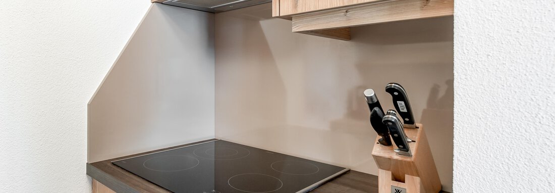 A modern kitchen unit with wooden fronts and a ceramic cooktop. On the countertop, there is a knife block with several knives. - Villa Castelbell - Sölden
