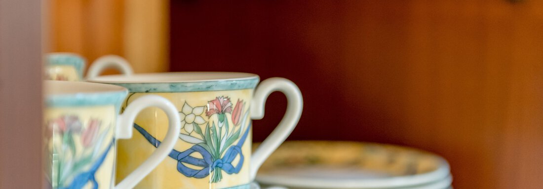 Stacked cups with a colorful flower design and some plates in the background. The cups are placed on a wooden shelf. - Villa Castelbell - Sölden
