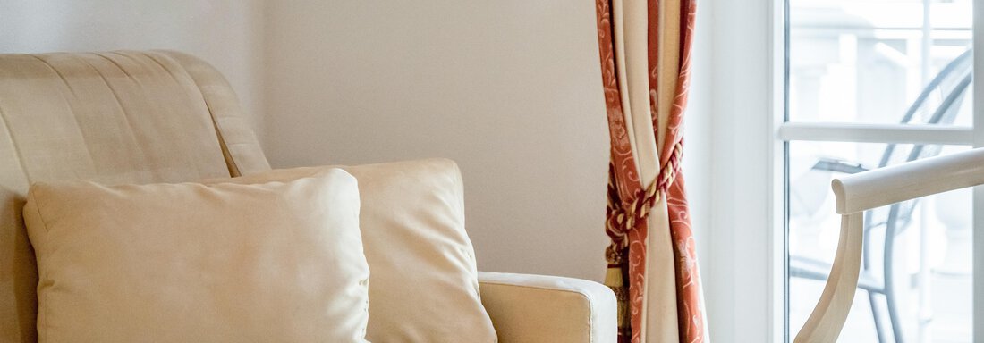 A cozy living room with a beige sofa and cushions.  
Curtains hang next to a window that lets in daylight. - Villa Castelbell - Sölden