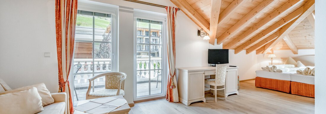 A bright room with a wooden ceiling and large windows. There is a sofa, a table, and a television. - Villa Castelbell - Sölden