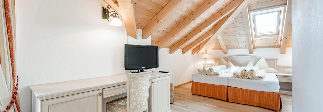 A bright room with slanted ceilings and wooden surfaces. There is a bed, a desk, and a television. - Villa Castelbell - Sölden