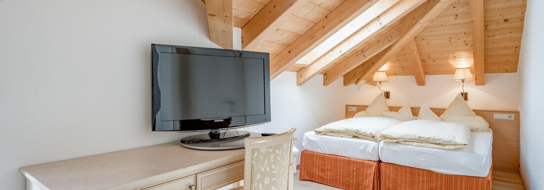 A cozy room with sloped ceilings and wooden beams. There is a double bed, a table with a chair, and a television. - Villa Castelbell - Sölden
