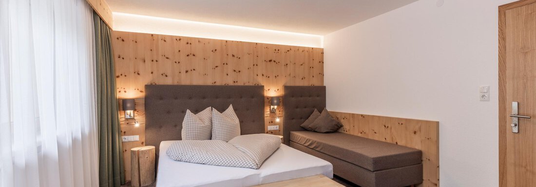 A modern hotel room with a double bed and a cozy seating area. The walls are made of light wood, and large windows provide plenty of daylight. - Vizensn Living - Sölden