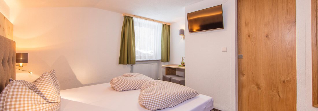 A cozy hotel room with two beds, a desk, and a television. The walls are bright and the window lets in plenty of daylight. - Vizensn Living - Sölden