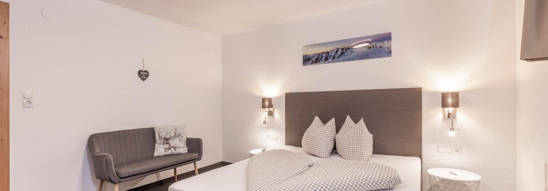 A modern bedroom with a double bed and a cozy armchair. The walls are light and simple, and there is decorative wall art. - Vizensn Living - Sölden