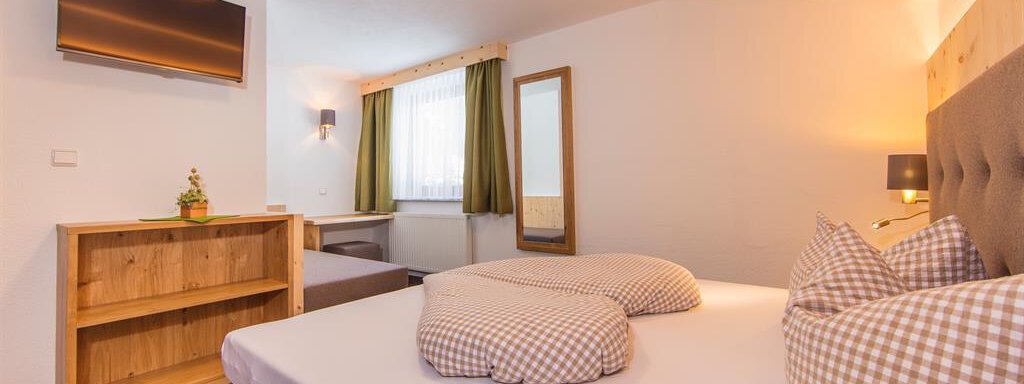 A cozy hotel room with a double bed and checked pillows. There is a flat-screen TV and a wooden wardrobe, as well as a large window front with green curtains. - Vizensn Living - Sölden
