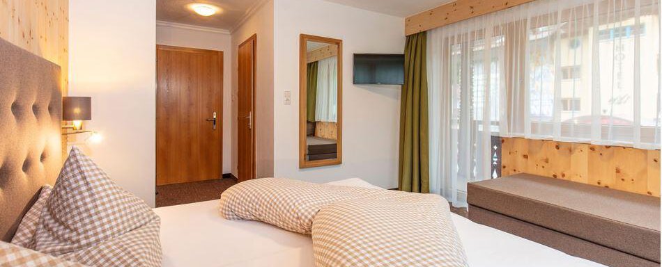 A cozy bedroom with a large bed and checked pillows. There is a wardrobe, a mirror, and plenty of daylight coming through the windows. - Vizensn Living - Sölden