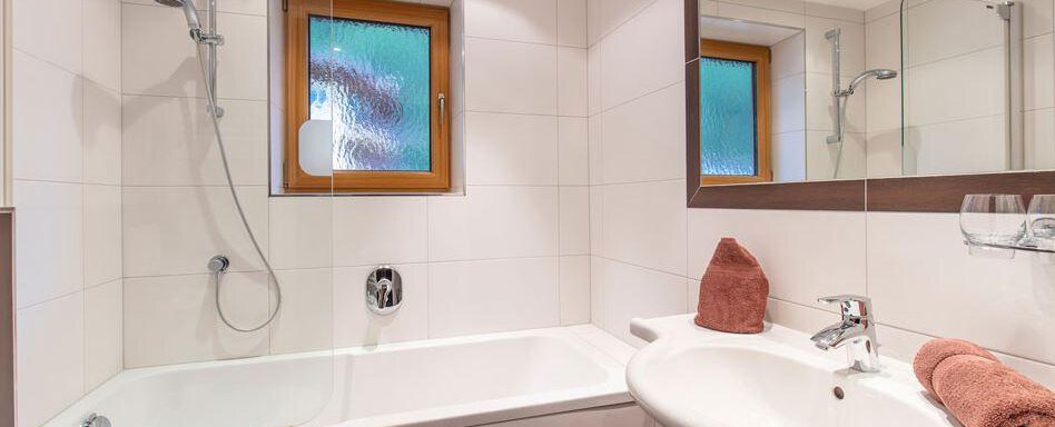 A modern bathroom with a bathtub and sink. The room is bright and friendly, with a window and a large mirror. - Vizensn Living - Sölden
