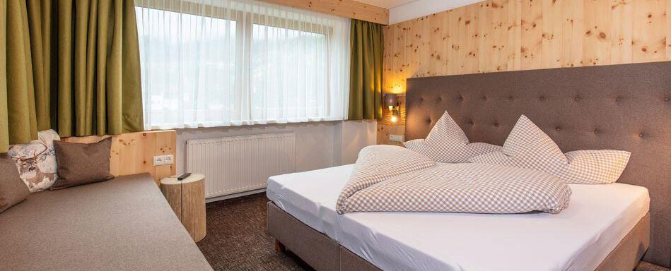 A modern bedroom with a double bed and a cozy sofa. The walls are made of wood and large windows provide plenty of daylight. - Vizensn Living - Sölden