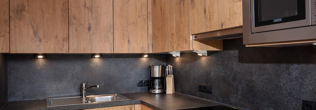 A modern kitchen with wooden fronts and a dark countertop. It features a sink, a cooking area, and modern kitchen appliances. - Vizensn Living - Sölden