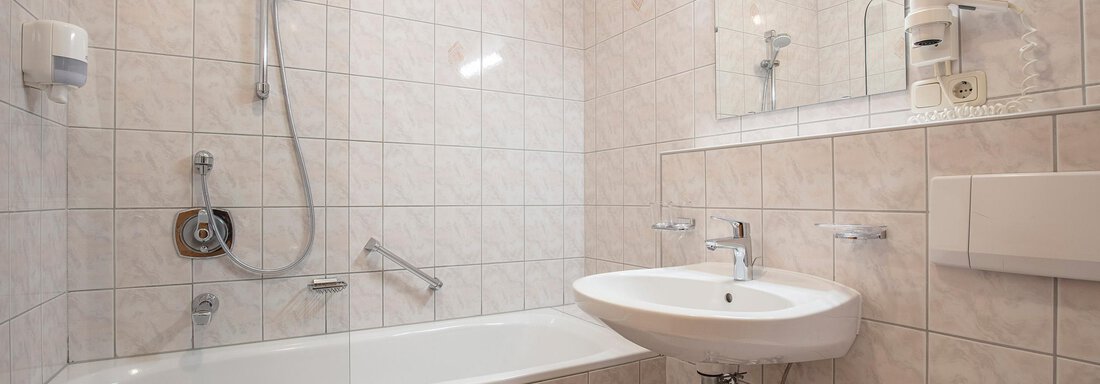 A bright bathroom with a bathtub, a sink, and a mirror. The walls are tiled with white tiles. - Vizensn Living - Sölden