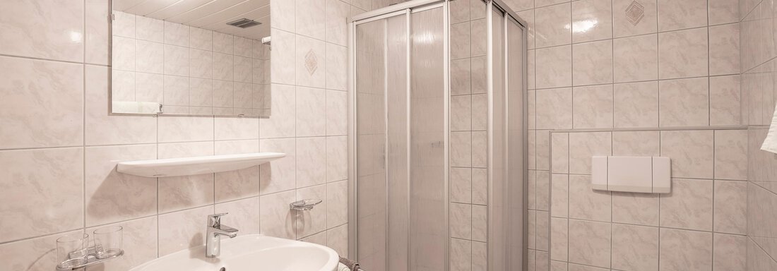 A modern bathroom with a shower, a sink, and a toilet. The walls are tiled white, and there is a large mirror. - Vizensn Living - Sölden