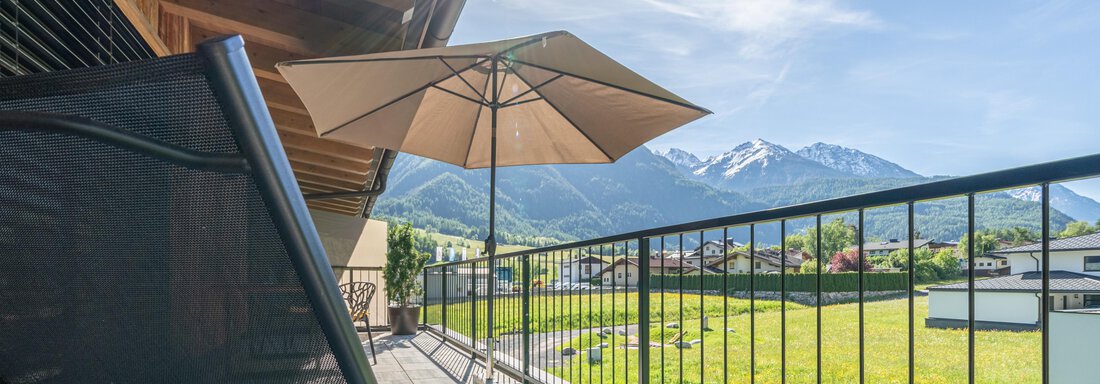 Ein Balkon mit einem Liegestuhl und einem Sonnenschirm. Im Hintergrund sind die Berge und ein klarer blauer Himmel zu sehen. - Apart Dorhoam - Umhausen