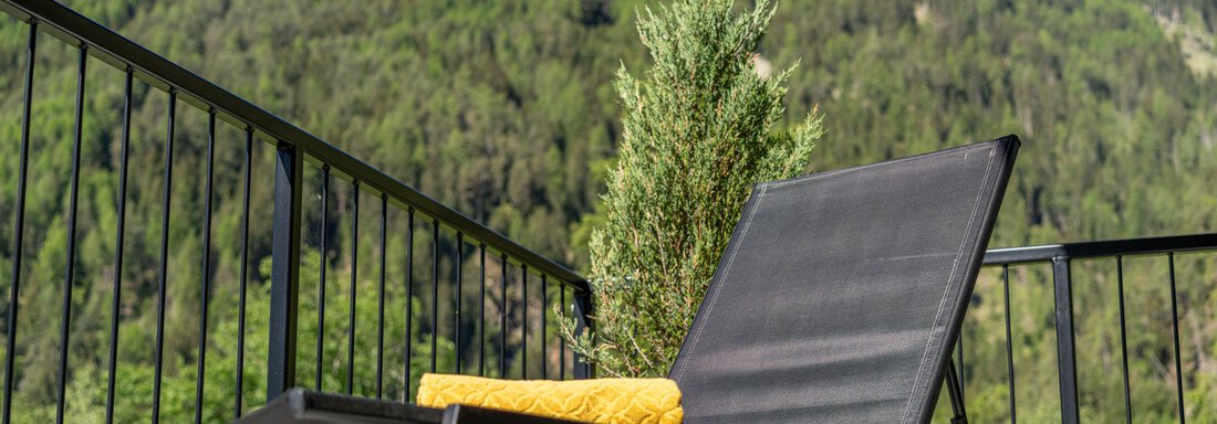 Ein bequemer Liegestuhl auf einem Balkon mit Blick auf eine grüne, bewaldete Landschaft und schneebedeckte Berge im Hintergrund. Neben dem Stuhl steht ein Topf mit einer Pflanze und ein gelbes Kissen. - Apart Dorhoam - Umhausen