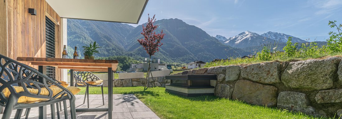 Eine Terrasse mit einem Holzschreibtisch und Stühlen. Im Hintergrund sind beeindruckende Berge und ein blauer Himmel zu sehen. - Apart Dorhoam - Umhausen