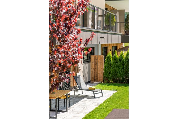 Ein gepflegter Garten mit einer Liege und einem Holztisch. Im Hintergrund sieht man moderne Gebäude und einen blühenden Baum. - Apart Dorhoam - Umhausen