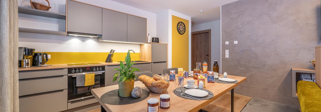 Eine moderne Küche mit grauen Schränken und gelben Akzenten. Ein schön gedeckter Tisch mit Snacks und Getränken lädt zum Essen ein. - Apart Dorhoam - Umhausen
