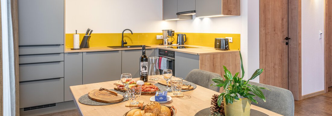 Eine moderne Küche mit grauen Schränken und einem Holztisch. Der Tisch ist liebevoll gedeckt mit Essen, Getränken und Pflanzen. - Apart Dorhoam - Umhausen
