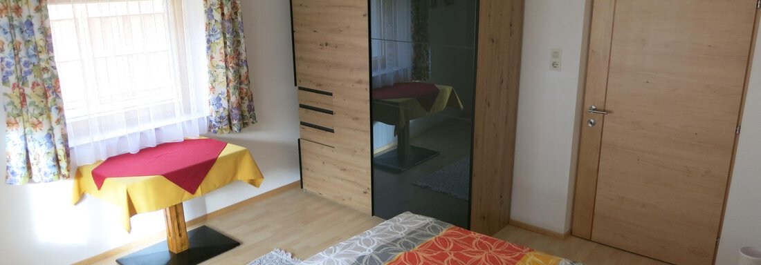 A bright bedroom with a large bed and colorful bedspreads. Decorative curtains hang at the window, and a table stands in the corner. - Apart Martina - Umhausen