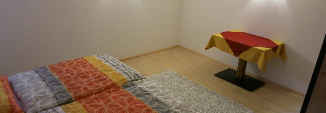 A simple room with a double bed and colorful bedspreads.  
On the right side, there is a table with a red tablecloth. - Apart Martina - Umhausen
