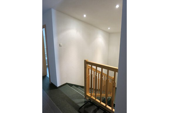 A bright stairwell with a wooden staircase and modern wall lights. The walls are plain and white, the floor is dark and elegant. - Apart Martina - Umhausen