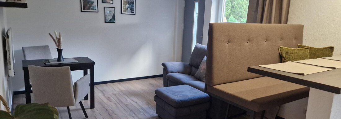 Ein gemütlicher Wohnraum mit einem Tisch und Stühlen, einer Couch und einem kleinen Hocker. An der Wand hängen Bilder, und das Licht kommt durch ein Fenster. - Apart Waldnest - Umhausen