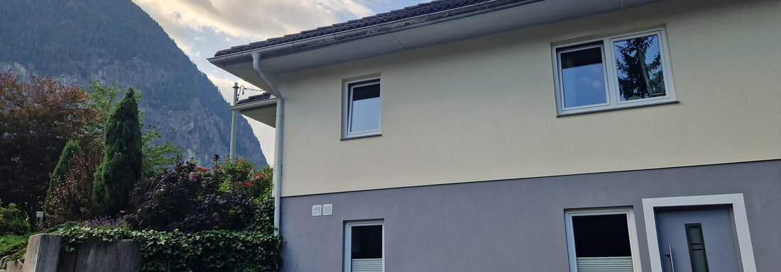 Ein modernes Haus mit einer grauen Fassade und großen Fenstern. Im Hintergrund sind Berge und ein bewölkter Himmel zu sehen. - Apart Waldnest - Umhausen
