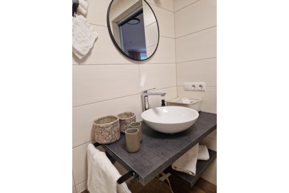 Ein modernes Waschbecken mit einer runden Spiegelfläche in einem stilvollen Badezimmer. Auf der Ablage stehen dekorative Behälter und ein Handtuch. - Apart Waldnest - Umhausen