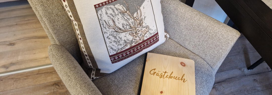 Ein gemütlicher Sessel mit einem dekorativen Kissen und einem Holzbuch mit der Aufschrift "Gästebuch". Der Boden ist aus Holz und der Raum wirkt einladend. - Apart Waldnest - Umhausen