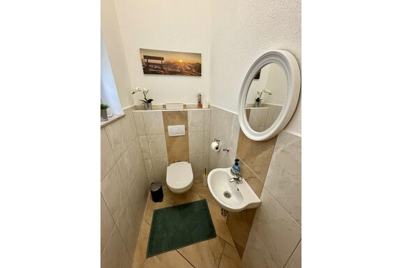 Ein kleines, modernes Badezimmer mit einer Toilette und einem Waschbecken. An der Wand hängt ein Spiegel und ein Bild mit Landschaftsmotiv. - Appartement Bergglück - Umhausen